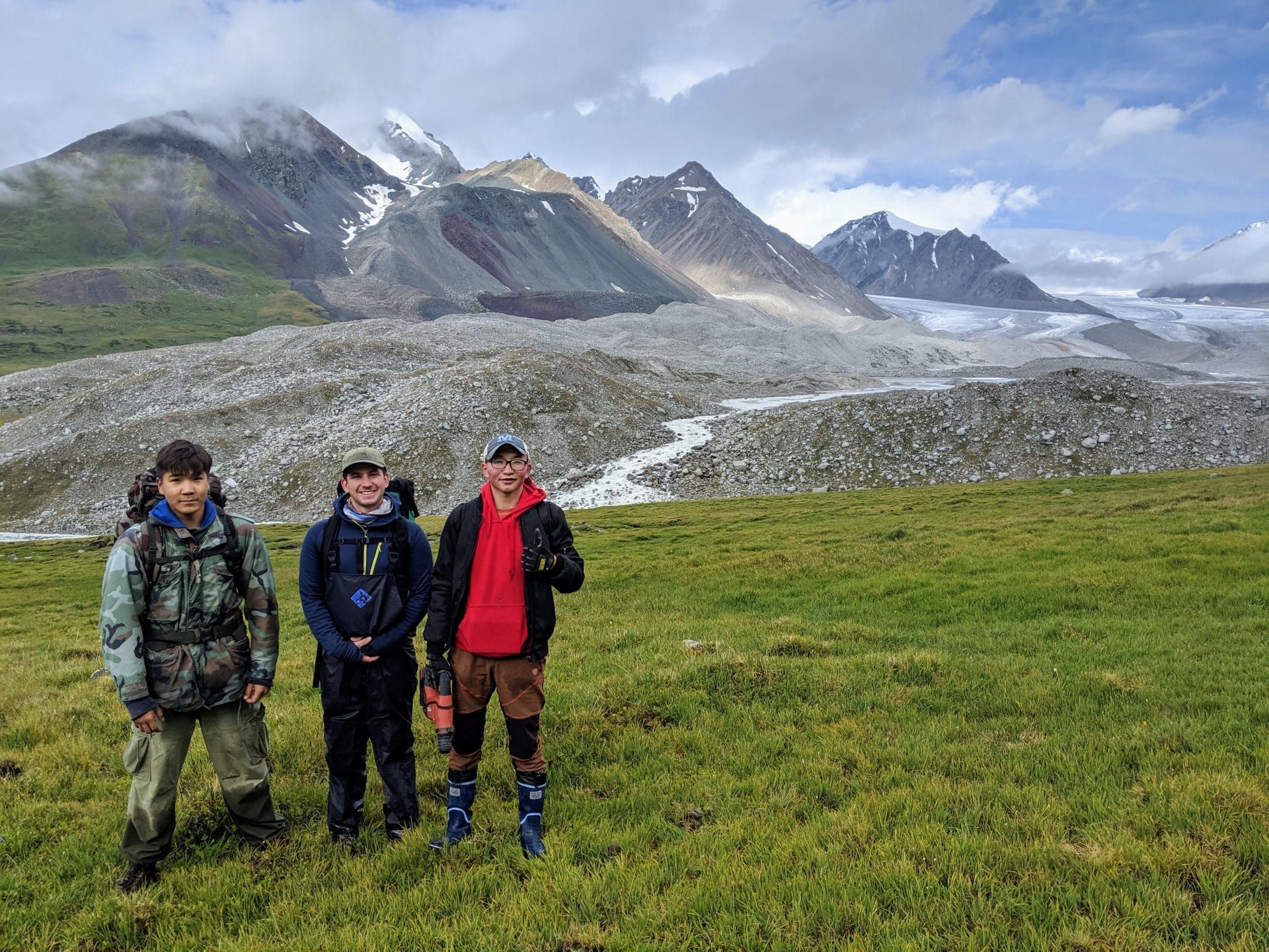 Team members Tumur Batbold, Peter Strand, and Oraza Seribol stand at the verdant edge of the Potanin glacier valley. The hills of tumbling rocks reveal young moraines.