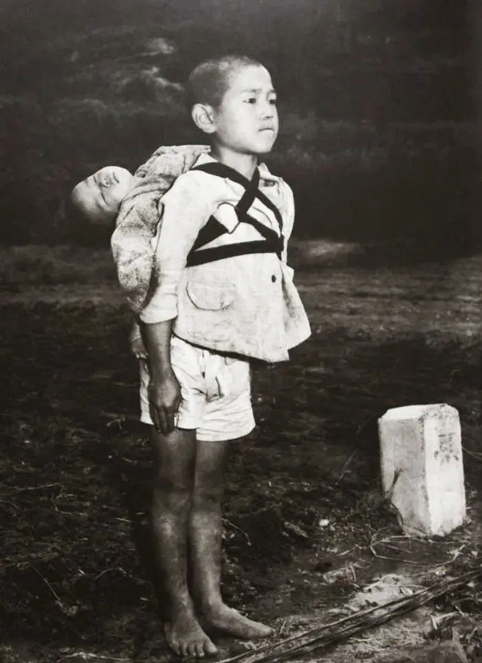 A barefoot boy waiting in line and staring ahead at a crematorium in Nagasaki, with his dead baby brother strapped to his back. Photo by US Marine photographer Joe O’Donnell