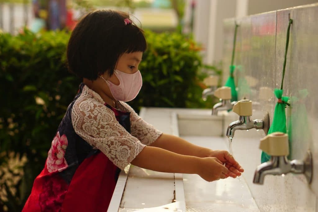 A girl washes her hands while wearing a mask. Credit: Pixabay.
