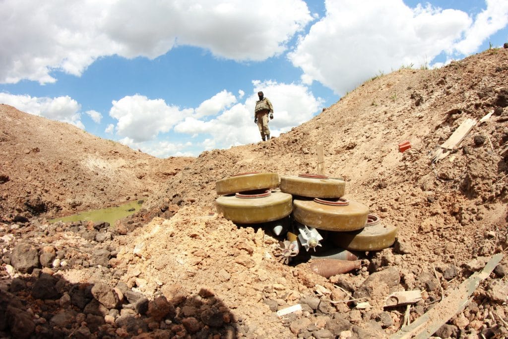 Soldier stands over land mines in front of partly cloudy sky.