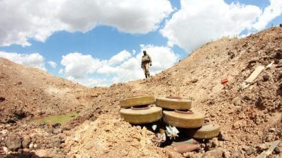 Soldier stands over land mines in front of partly cloudy sky.