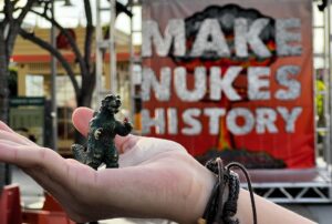 toy Godzilla held in a hand in front of Make Nukes History sign