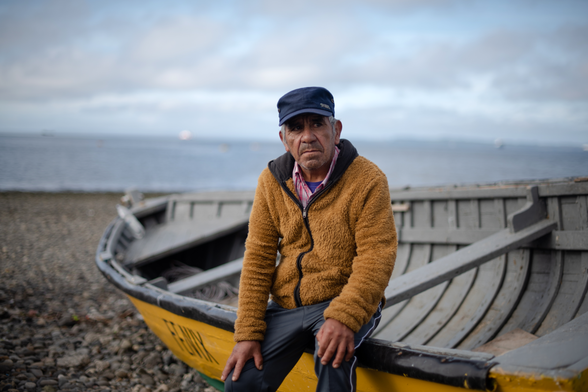 A man sits on the side of a fishing boat in a yellow jacket and hat. The ocean is behind him.