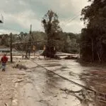 A man walks towards a flooded river, across a mud-strewn road.