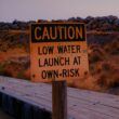 Sign reading "Caution Low Water Launch At Own Risk" at Great Salt Lake marina during a drought in front of naturally-colored grasses that look like fire.