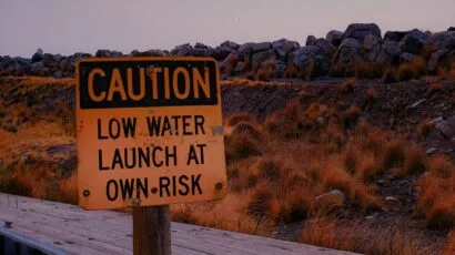 Sign reading "Caution Low Water Launch At Own Risk" at Great Salt Lake marina during a drought in front of naturally-colored grasses that look like fire.