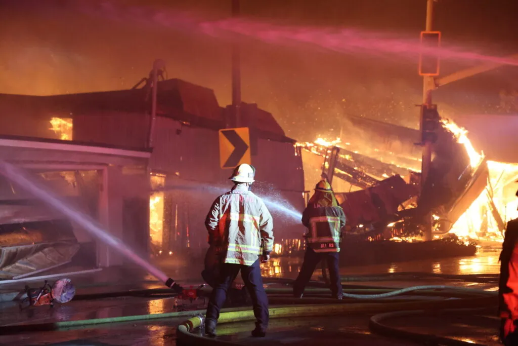 two firefighters spraying burning buildings with hoses with a destroyed building in flames in the background