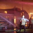 two firefighters spraying burning buildings with hoses with a destroyed building in flames in the background