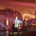 two firefighters spraying burning buildings with hoses with a destroyed building in flames in the background