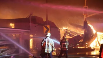 two firefighters spraying burning buildings with hoses with a destroyed building in flames in the background