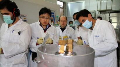 A group of scientists in lab coats and masks observe and take notes around a metal scientific apparatus in a laboratory setting.