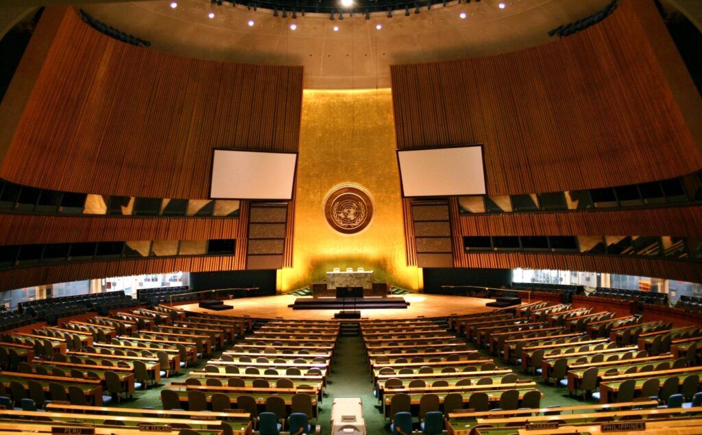 Large empty assembly hall with rows of seats a central podium and the United Nations emblem displayed on a golden wall in the background