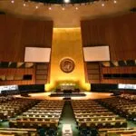 Large empty assembly hall with rows of seats, a central podium, and the United Nations emblem displayed on a golden wall in the background.