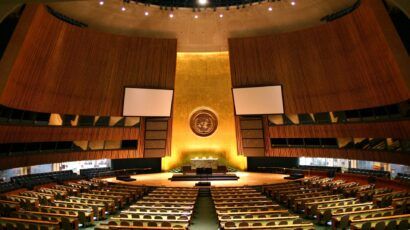 Large empty assembly hall with rows of seats, a central podium, and the United Nations emblem displayed on a golden wall in the background.