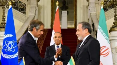 Two officials shake hands at a formal event, with IAEA and Iranian flags on the table, and three national flags in the background. A third person stands between them, clapping.
