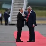 Two men in suits greet each other on a red carpet at an airport, with an airplane and several people standing in the background.