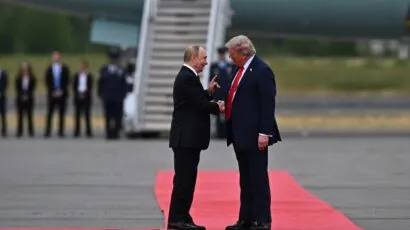 Two men in suits greet each other on a red carpet at an airport, with an airplane and several people standing in the background.
