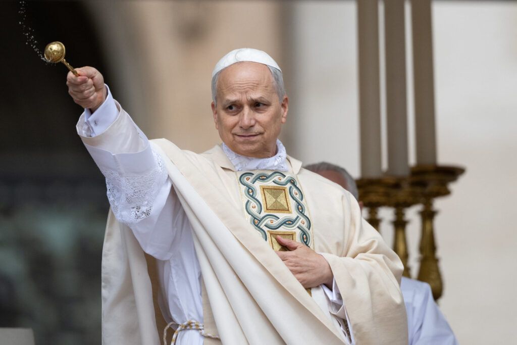 A Catholic priest in white liturgical vestments sprinkles holy water during a religious ceremony, with large candles in the background.