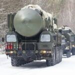Military trucks carrying large missile systems drive on a snow-covered road through a forested area.