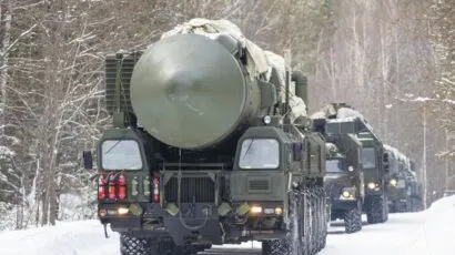 Military trucks carrying large missile systems drive on a snow-covered road through a forested area.
