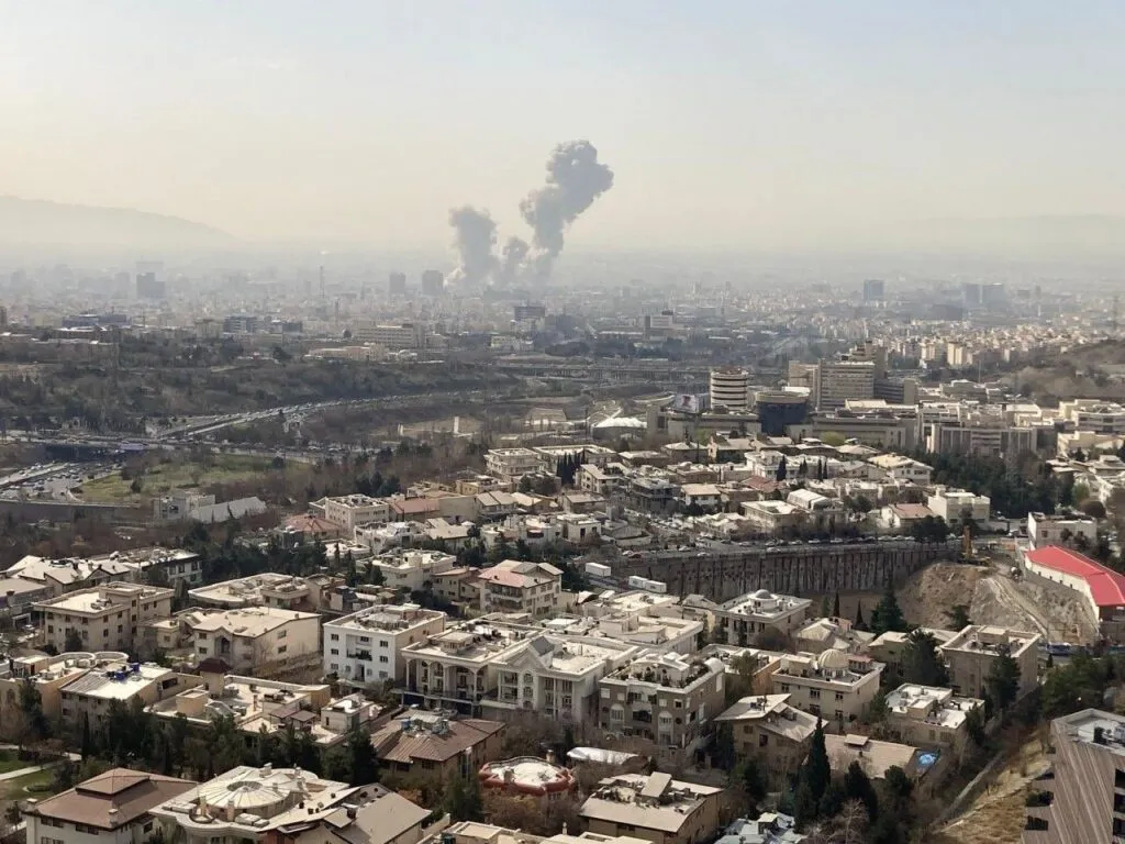 A cityscape with a plume of smoke rising in the distance under a hazy sky, indicating an explosion or fire in the urban area in Iran.