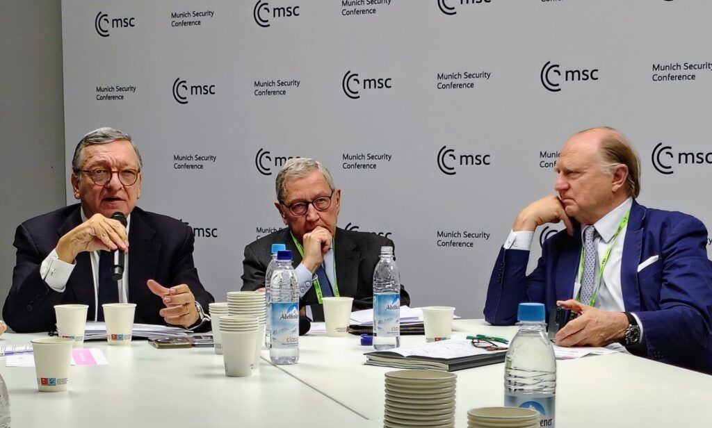 Three men in suits sit at a conference table with drinks and documents, speaking and listening during the Munich Security Conference.