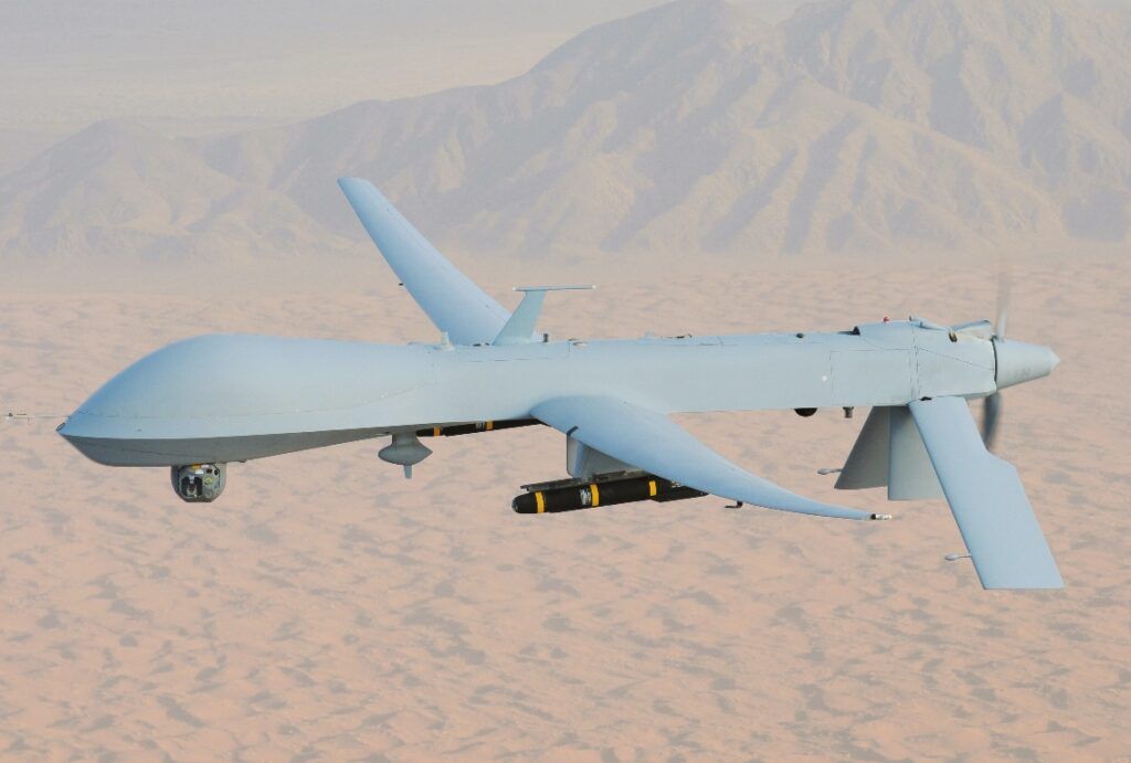 A white MQ-1 Predator drone flying over land with mountains in background.