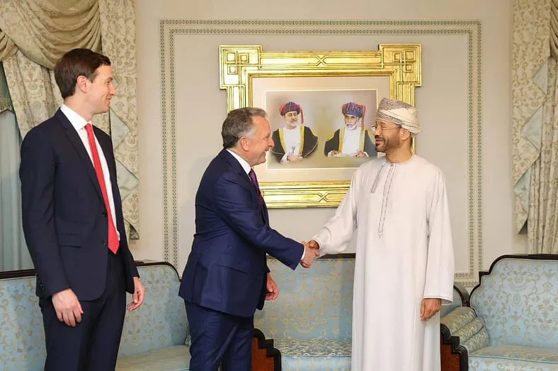 Three men meet in a formal room; two men in suits shake hands while a man in traditional Omani attire stands opposite them. A framed portrait hangs on the wall behind them.