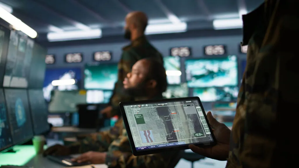Army professional holding a screen and employing AI tech to improve military combat systems and missiles monitoring. In the background are screens and two other military professionals.