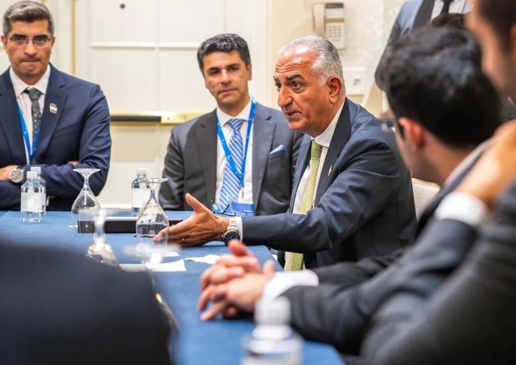 A group of men in business attire sit around a conference table, engaged in discussion; one man gestures while speaking.