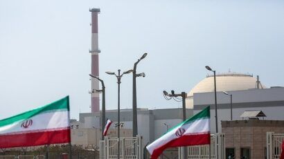 Iranian flags wave in front of a nuclear power plant complex with a dome-shaped reactor building and a tall red-and-white smokestack in the background.