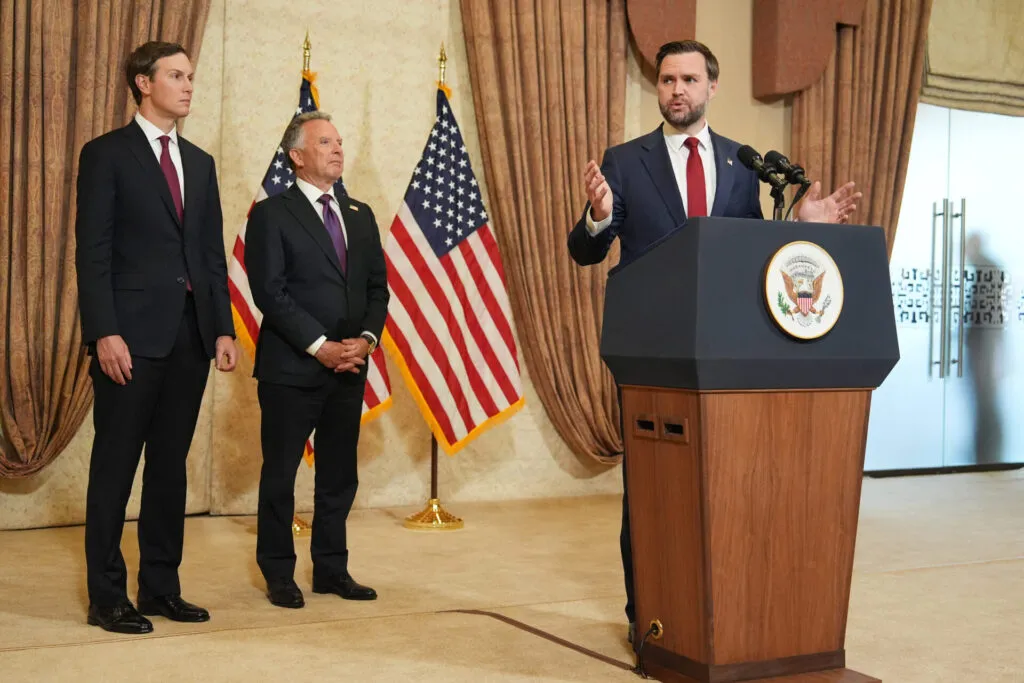 Three men in suits stand in front of two US flags; one speaks at a podium with the Vice Presidential seal, while the other two listen.