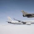 A French military jet flies above a French Air Force refueling aircraft in midair against a clear sky.