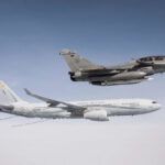 A French military jet flies above a French Air Force refueling aircraft in midair against a clear sky.
