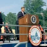 A man in academic regalia speaks at a podium with the presidential seal during an outdoor ceremony at The American University, with people seated behind him.