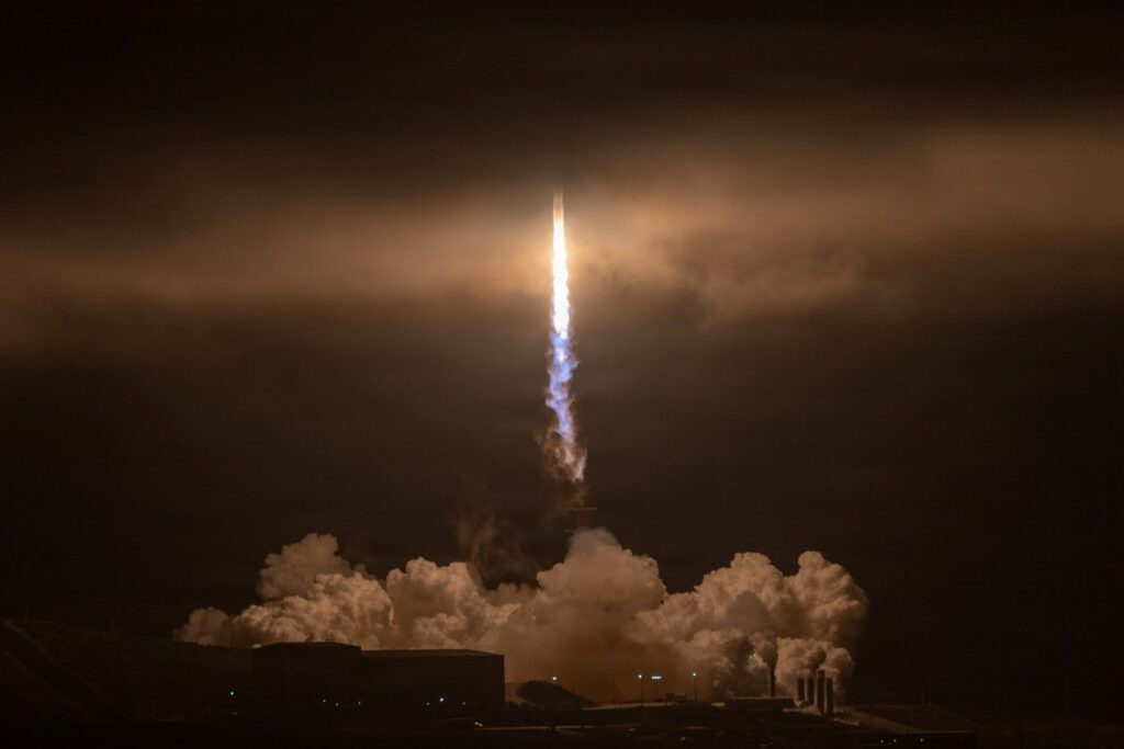 A rocket launches at night, rising vertically through clouds with bright light and smoke trailing behind, silhouetting buildings and structures on the ground.