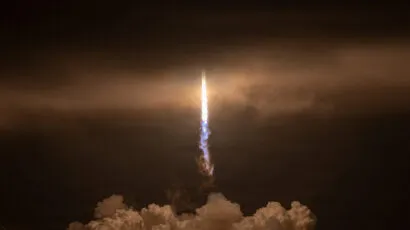 A rocket launches at night, rising vertically through clouds with bright light and smoke trailing behind, silhouetting buildings and structures on the ground.