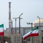 Iranian flags wave in front of a nuclear power plant complex with a dome-shaped reactor building and a tall red-and-white smokestack in the background.