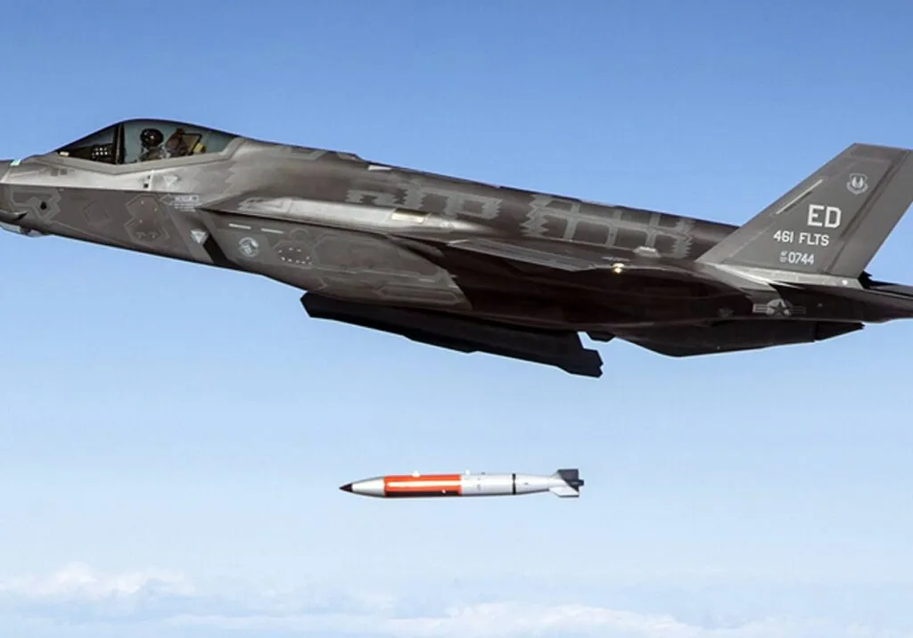 An F-35A fighter jet releases a B61-12 dummy nuclear bomb mid-flight against a clear blue sky.