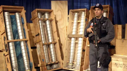 A security officer stands armed next to large metal pipes packed in wooden crates, displayed upright against a blue curtain backdrop.