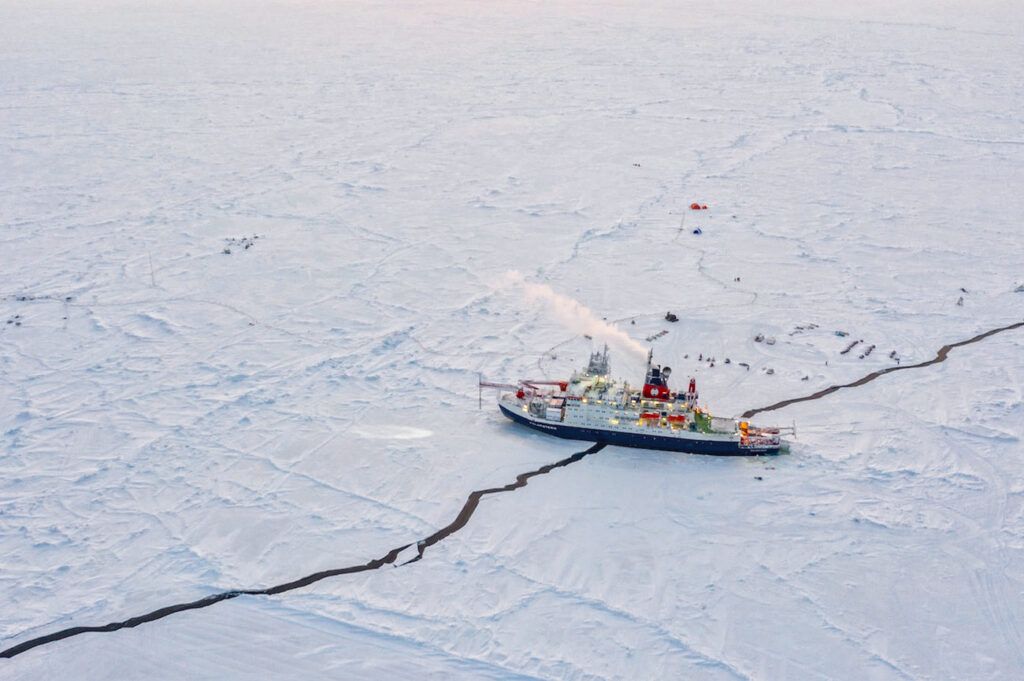 An icebreaker ship navigates through a cracked, frozen sea with scattered equipment and tents visible on the ice.