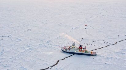 An icebreaker ship navigates through a cracked, frozen sea with scattered equipment and tents visible on the ice.