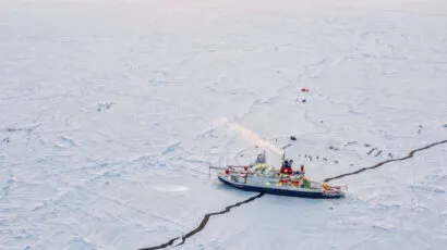 An icebreaker ship navigates through a cracked, frozen sea with scattered equipment and tents visible on the ice.