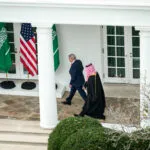 Two men walk outside a white building past American and Saudi Arabian flags; one wears a suit and the other wears traditional Middle Eastern attire.