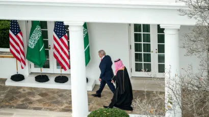 Two men walk outside a white building past American and Saudi Arabian flags; one wears a suit and the other wears traditional Middle Eastern attire.