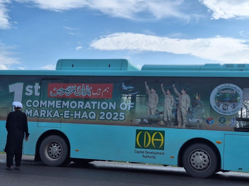 A person stands near a blue bus displaying a banner for the 1st Commemoration of Marka-e-Haq 2025, with images of soldiers and the Capital Development Authority logo.