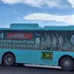 A person stands near a blue bus displaying a banner for the 1st Commemoration of Marka-e-Haq 2025, with images of soldiers and the Capital Development Authority logo.