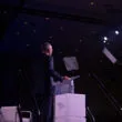 A man in a suit stands at a podium on stage, facing an audience, with two teleprompters in front of him and stage lights shining above.