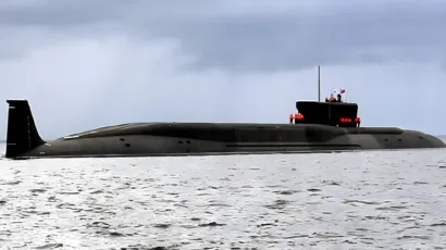 A large, black military submarine is partially submerged and moving through calm waters under a cloudy sky.