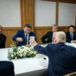 Leaders from the United States and China sit around a conference table with documents, flowers, and flags, engaging in a formal discussion.